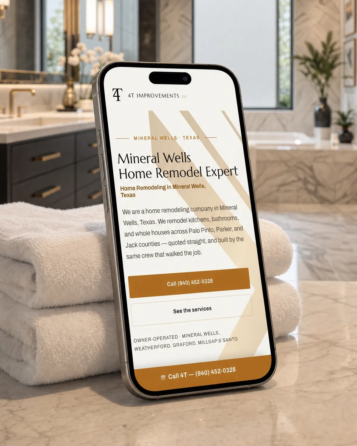 4T Improvements home page on a phone standing on the marble counter of a finished bathroom remodel, folded towels beside it.
