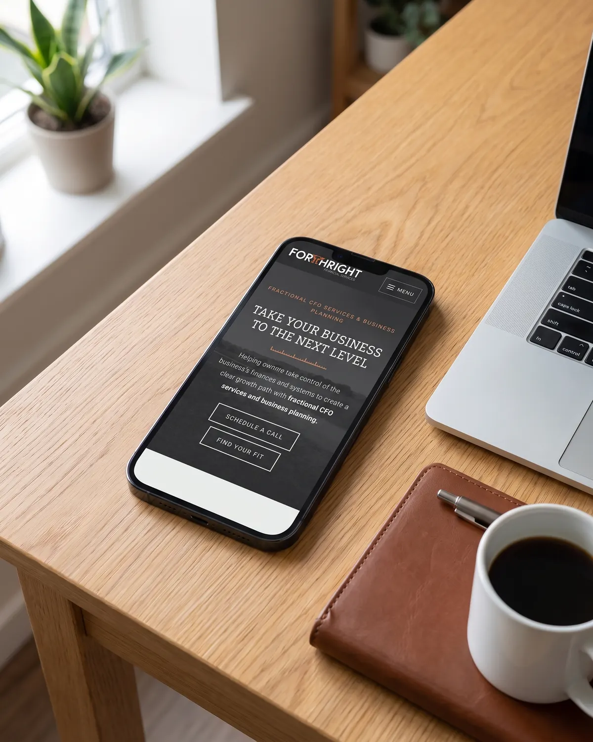 Forthright Financial Services home page on a phone lying on an oak desk beside a laptop, a leather notebook and a cup of coffee.
