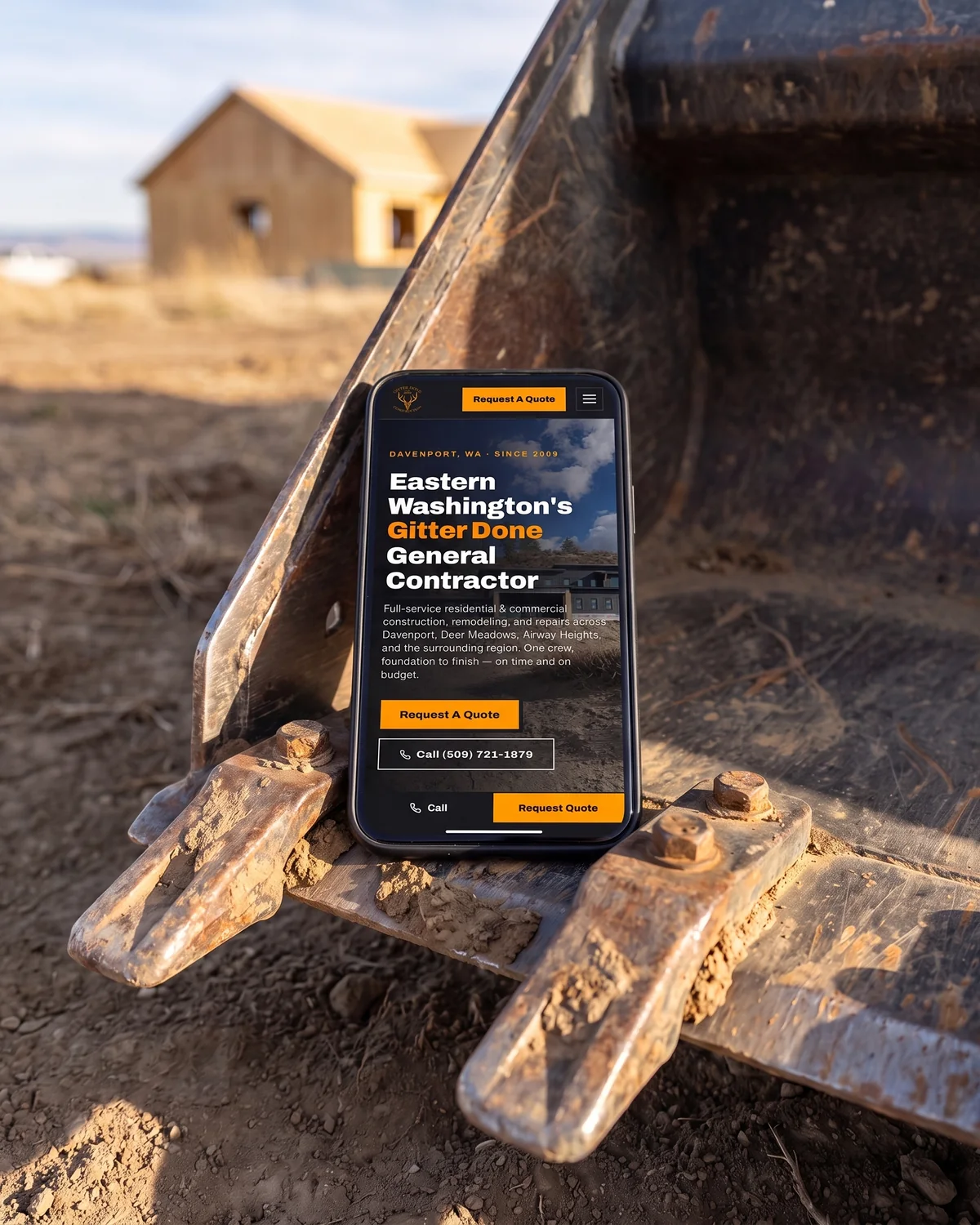 Gitter Done Construction home page on a phone lying across the teeth of a skid steer bucket on a jobsite.