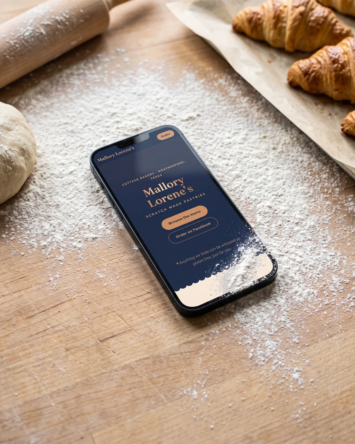 Mallory Lorene’s bakery home page on a phone lying in sifted flour on a butcher-block counter.