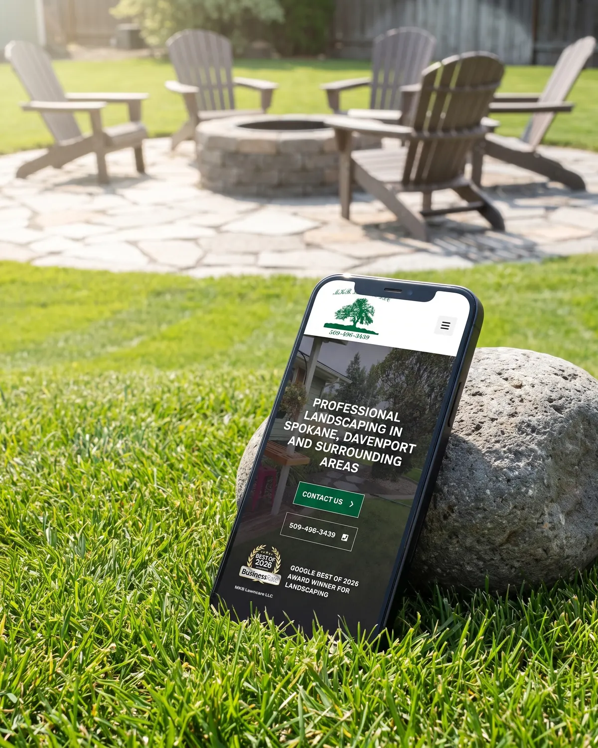 MKB Lawn Care home page on a phone leaning against a rock at the edge of a fresh-cut lawn, with a patio and fire pit behind.